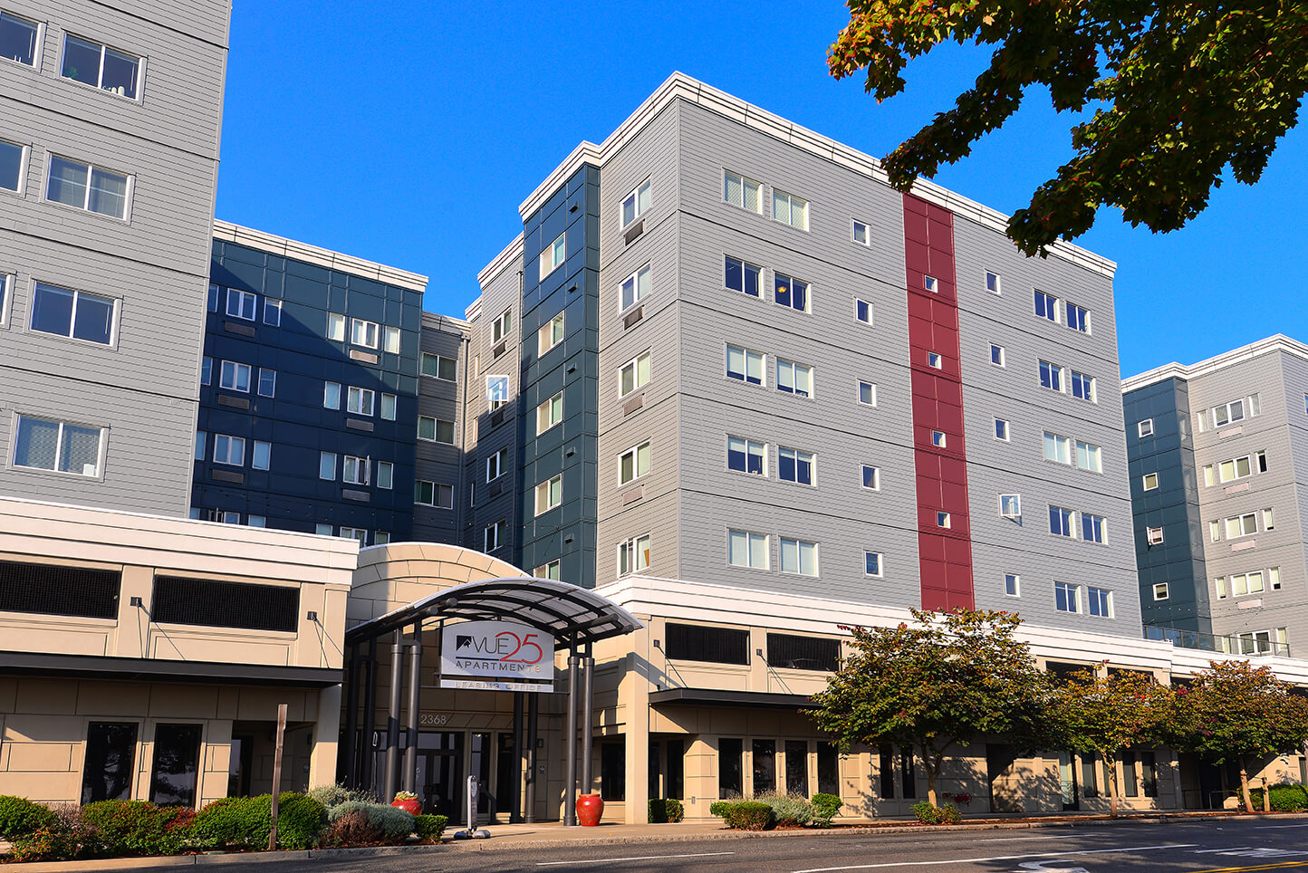 A modern, multi-story apartment building with gray and red accents, large windows, and trees in front. The entrance features a covered walkway and a sign reading Plaza 525.